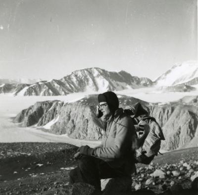 A man wearing a coat, backpack, and warm hat knelt on the ground on top of a rocky hill surrounded by other rocky, snow-covered hills