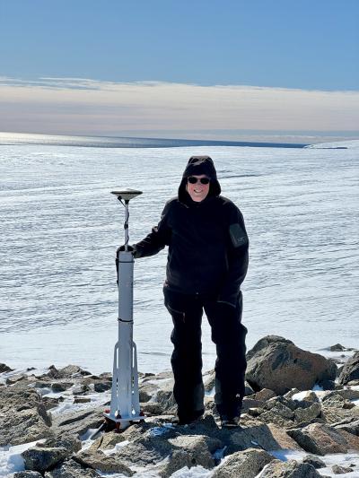 A person wearing a black ski jacket and sunglasses standing next to the GNSS antenna, with a vista of the ice-covered Sea behind.