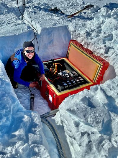 A person in winter gear and sunglasses sitting in a dug up square ditch next to a rectangular red case containing equipment, surrounded by snow.