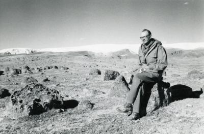 Black and white photo of man with glasses and long jacket sitting on a rock in a rocky field with snow and ice in a distance