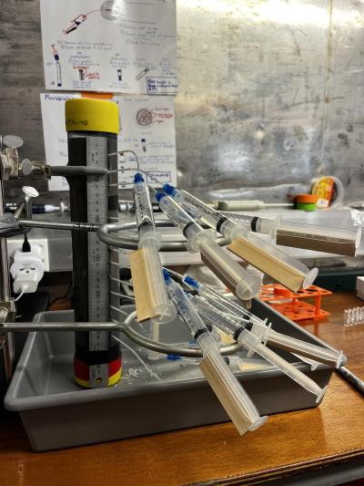A number of syringes with clear liquid attached via clear tubes to a cylinder column with a ruler attached to it in a short plastic tub on table in a lab