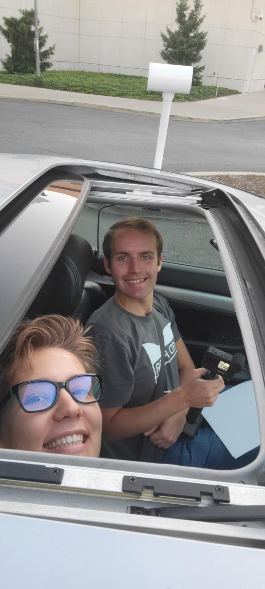 Geddy Davis with short light hair smiling wearing a gray T-shirt with Byrd and image in white further from the camera and only Karina's Peggau smiling face showing with short hair and black frame glasses in forefront both looking up though the open sunroof of a light color car parked in a lot with asphalt and sidewalk and grass and trees in background  and Apparatus sticking up from the car