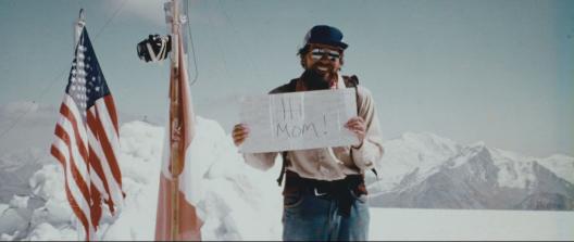 Lonnie Thompson standing outdoors in snow on a sunny day next to an American flag, holding a sign that says Hi mom.