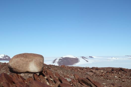 a brown rocky ground with a boulder in the foreground and snow covered landscape in the background.