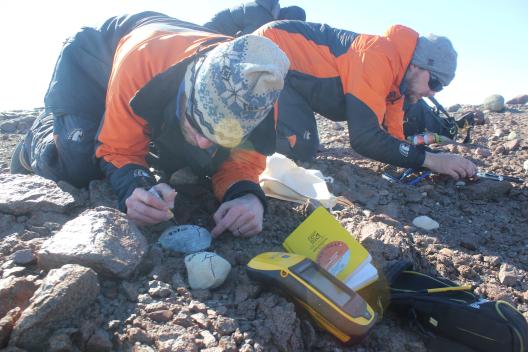 Two men in winter jackets, sunglasses and hats kneeling on the dirt ground  writing on rocks with markers