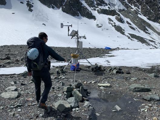 Someone walking in a rocky terrain toward field equipment with some snow on the ground.