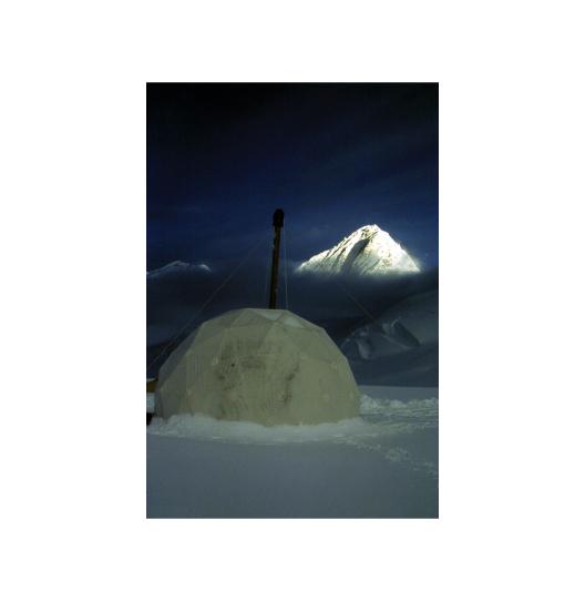 A white dome tent on the snowy ground and a white snow capped mountain at a distance under dark blue skies.