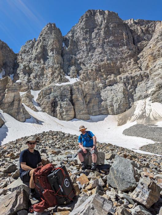 Two people sitting on rocks in the mountainous field surrounded by rocks and snow.