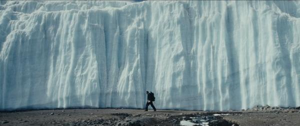 A man with a backpack walking on dirt surface along a wall of ice many times higher than him  during daylight.