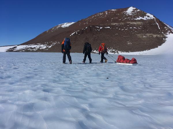 Snow covered ground with a  rock mountain and three people walking away from the camera. one pulling a red sled behind him