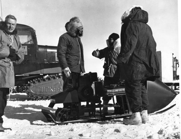 Black and white of four people standing, three talking, one looking at the camera,  in winter gear outdoors next to a snow mobile and snow tractor.