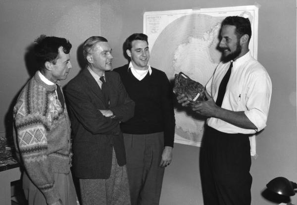 Black and white image of a group of four men standing in a room, one of them showing the others a rock he is holding in his hands with the map of Antarctica in the background.