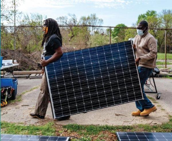 Two men wearing masks carrying a large solar panel outdoors