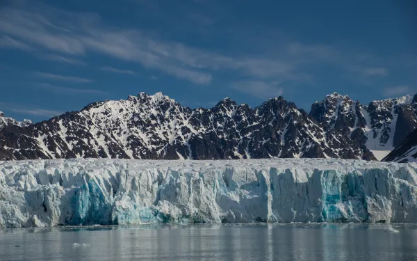 A pano of a lake and glacier and a rocky mountain with snow streaks under blue skies and light, white clouds