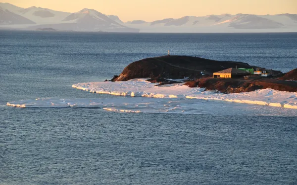 A view of the somewhat frozen lake and a house on a bedrock in a distance with snow covered mountains  in the background under cream colored sky,