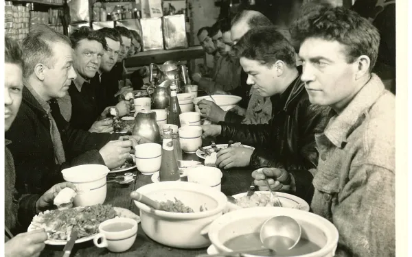 12 collective men seated before a buffet streching out across a wooden bench. A cutoff menu in the top right corner indicates the setting as a restaraunt.