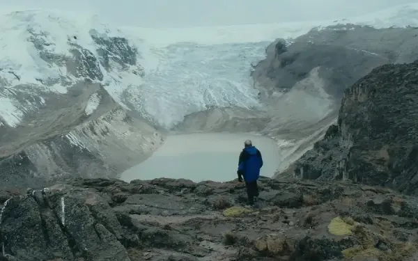 Person in blue jacket standing on rocky ground, observing a glacier and a glacial lake in a vast, mountainous landscape.
