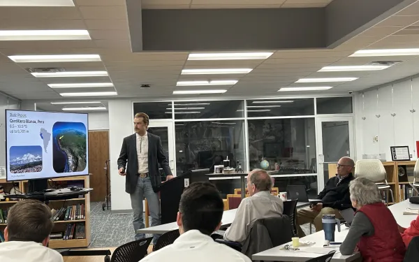 A presentation with an audience listening to a speaker standing next to a screen.