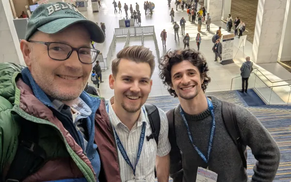Three smiling people pose for a selfie in a spacious convention center lobby.