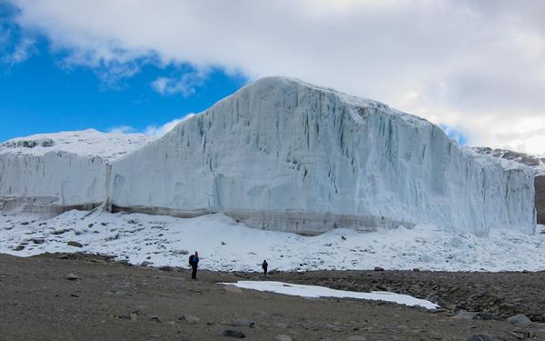 A large glacier. Walking towards its base are two figures.