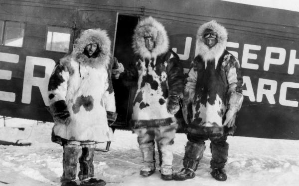 Three people pose for a photo in front of a plane in the arctic. They are wearing cold weather gear and large fur lined coats 