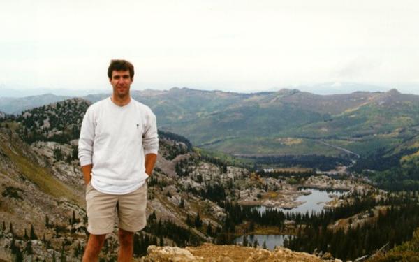 A white man in tan shorts and a white shirt with long sleeves rolled half way up his arm. He is standing in front of mountain and valley landscape.