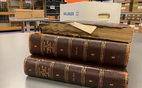A stack of three brown books with worn down covers. They are placed on a plain table in an archival room. 