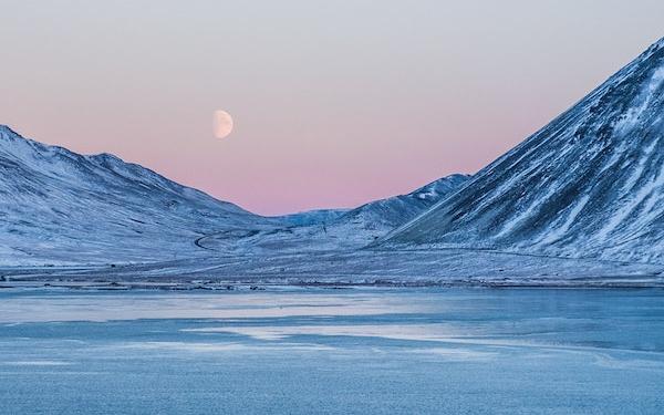 Frozen lake in Iceland surrounded by snowy mountains that border either side of the image like a bowl, framing the moon and the pink sunset
