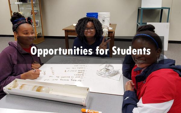 Three smiling, young, black students sitting at a table with a poster board and crafting materials around them. Overtop is the text "opportunities for students"