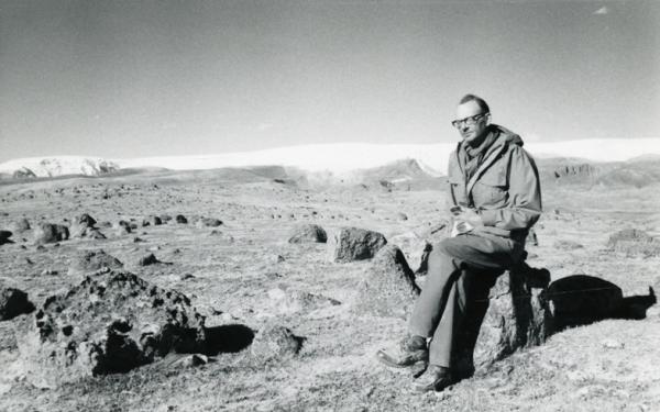 Black and white photo of man with glasses and long jacket sitting on a rock  in a rocky field with snow and ice in a distance
