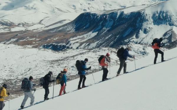 A group of people with backpacks climbing up a snow covered mountain, walking in a row,