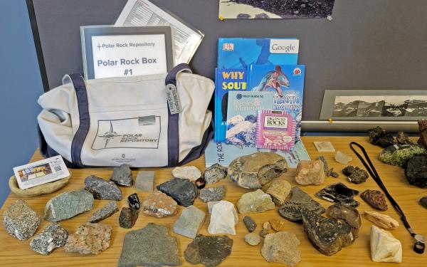A canvas bag on a wooden table with a Polar rock Repository logo on it and a notebook with the same title peeking out from it. On the desk there are rocks laid out and books next to the bag.