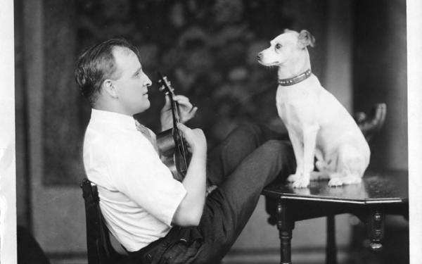 A man sitting at a table with his legs stretched on the table, playing a ukulele with a dog facing him sitting on the table in front of him.