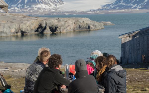 A small group of people sitting at a table outdoors under the sunlight, looking out at a body of water close by with snow covered mountains and bedrock with some gray clouds.