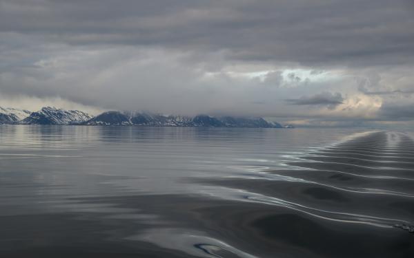 Body of water with a visible series of ripples and snow covered mountains to the left in a distance with gray and white cloud cover,.