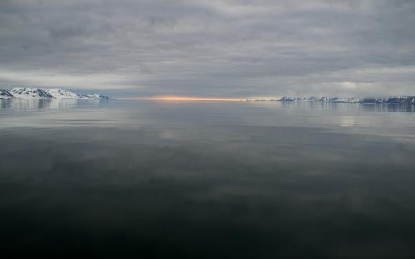 Body of water surrounded by snow covered mountains in a distance with sunlight in the horizon under gray cloud cover,