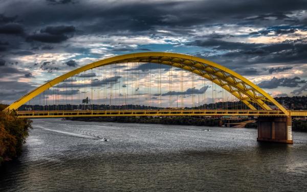 A yellow bridge over a river with a background of dark and white clouds  showing some blue sky.