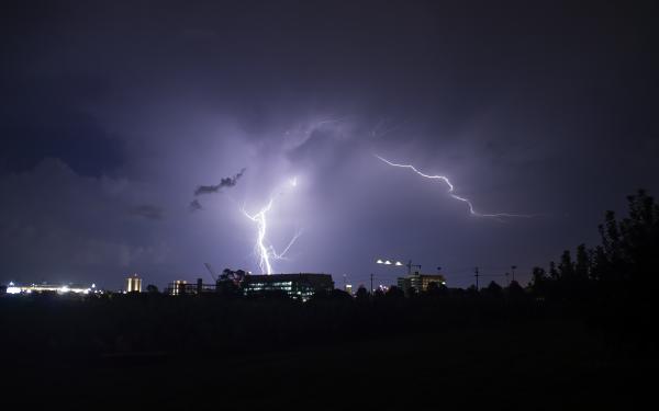 Bolts of lightning lighting up the night sky in the distance as a backdrop to taller buildings among tree lines.