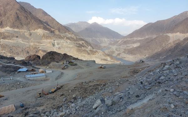 a low lying river in a rocky-dirt terrain in a valley surrounded by brown mountains with some structures, trucks and machinery under blue skies and white clouds.