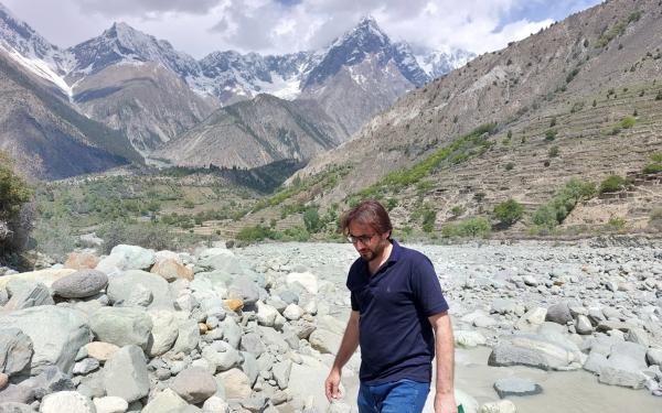 A man holding a green-covered notebook walking in a rocky river bed with a little stream flowing behind him and sparse tree covered hills and mountains in the background. 