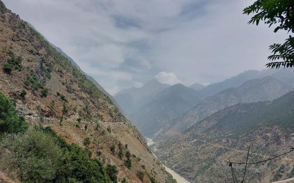 A dirt Mountain range covered sparsely with trees and a river flowing with light brown water below.