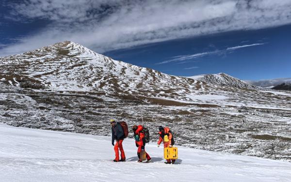 Three people walking up a snow covered hill, surrounded by mountains, with blue skies and white clouds..