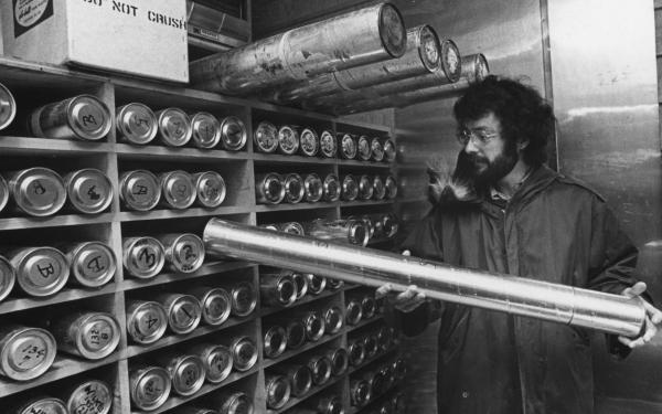 A black and white image of a bearded man with glasses in a winter coat holding a silver cylinder, against a shelf filled with cylinders along the wall.