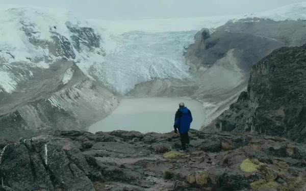 Person in blue jacket standing on rocky ground, observing a glacier and a glacial lake in a vast, mountainous landscape.