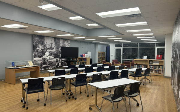 Interior of a modern library with tables and chairs arranged for reading or studying. The room features large murals depicting historical scenes, bookshelves with books, and a flat-screen TV on one wall.