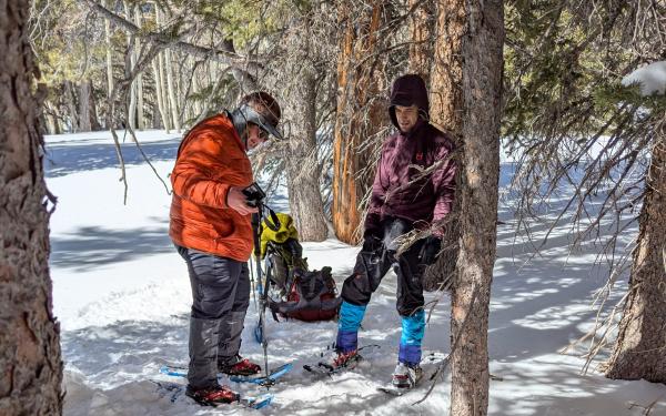 Two people with snowshoes adjusting their equipment in a snowy forest.