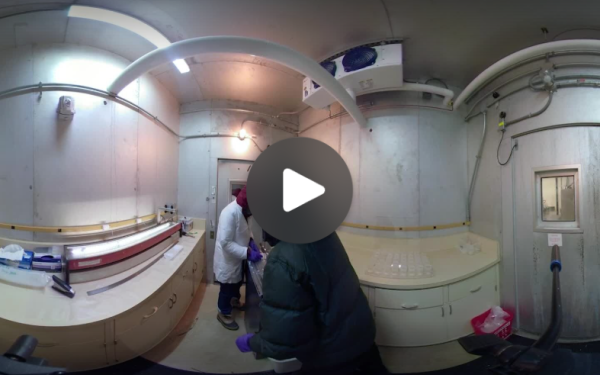 A 360-degree view of an indoor setting, with two individuals engaged in tasks, with a play icon in the center of the image.
