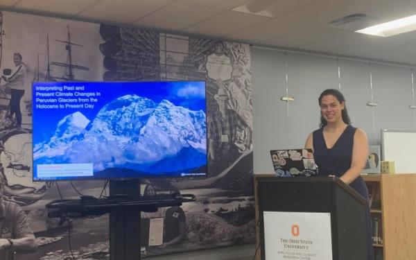 A person stands at a podium, next to a large display screen, giving a presentation and has a laptop in front of her.