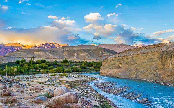 Scenic view of the Indus River flowing through a rugged landscape with lush green vegetation along the banks, under a vibrant sky at sunset.