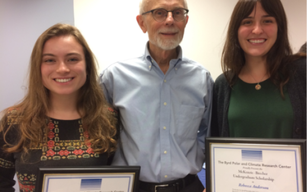Three individuals pose indoors against a plain background. The two individuals on either side are holding certificates.